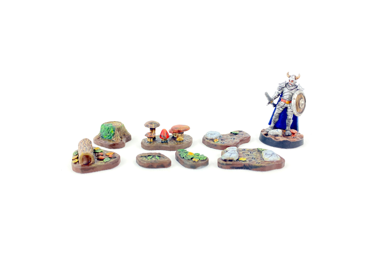 Alle sieben bemalten Waldboden-Bases aus dem Waldboden-Scatter-Set mit einer 28 mm Miniaturfigur (Krieger mit Schwert, Schild und Helm mit Hörnern) als Größenvergleich, zeigt Maßstab und Detailgrad für Tabletop-Spiele.