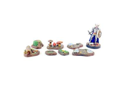 Alle sieben bemalten Waldboden-Bases aus dem Waldboden-Scatter-Set mit einer 28 mm Miniaturfigur (Krieger mit Schwert, Schild und Helm mit Hörnern) als Größenvergleich, zeigt Maßstab und Detailgrad für Tabletop-Spiele.